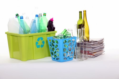 In the image, there is a bright green plastic recycling box with a blue recycling symbol on its side, filled with various used plastic bottles, some of which are partially crushed or empty, featuring different colors including blue, green, and clear. To the right of the green bin, there is a smaller blue plastic basket containing more empty plastic bottles, some with caps, and a few appear to be squeezed or crumpled. Adjacent to these containers, a neat stack of newspapers or magazines is visible, with the paper edges slightly curled and overlapping, placed on a flat surface. Next to the newspapers, several glass bottles of varying shapes and sizes are arranged, including slender, elongated bottles with coloured glass in shades of yellow and green, along with clear glass bottles, some with their caps or stoppers still attached. The background is plain white, with soft, even lighting that highlights the details and textures of the waste materials, suggesting a clean indoor setting suitable for a professional rubbish removal service in Northolt or the surrounding area.
