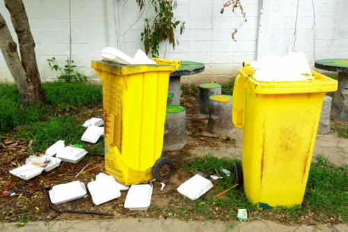 Two yellow wheeled waste bins positioned on a paved outdoor area adjacent to a white wooden fence with visible horizontal panels, surrounded by scattered white foam food containers and some debris on the grass and ground. The bins are made of durable plastic with textured surfaces and are partially open, revealing white trash bags inside. In the background, there are small round tables and stools with brightly painted surfaces, along with some green foliage and a weathered tree trunk on the left side, indicating a small communal or outdoor seating area. The scene captures a typical rubbish disposal point, with litter that appears to have been recently discarded, reflecting conditions suitable for professional waste removal services in Northolt or similar locations.