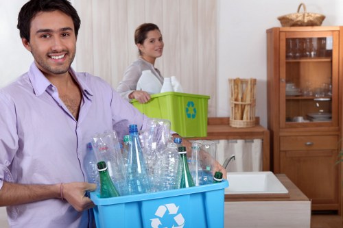 A man with dark hair, wearing a light purple shirt, is standing in a kitchen holding a large blue recycling bin filled with empty transparent plastic bottles, including green-tinted glass bottles, and a few aluminium cans, all clean and neatly arranged. Behind him, a woman with dark hair tied back, dressed in a light grey top, is leaning on a wooden kitchen counter next to a bright green recycling box with a blue recycling symbol, positioned near a white sink. The kitchen features wooden cabinets, one of which has glass doors displaying crockery, and a neutral-toned wall with vertical striped wallpaper. The scene is well-lit, likely from natural light, creating a clear and professional atmosphere related to waste management and recycling services, typical of commercial waste removal in Northolt.