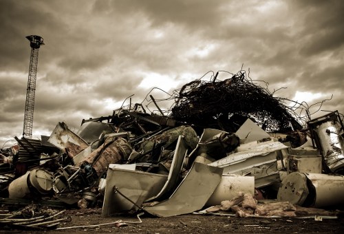 A large pile of scrap metal and discarded household appliances, including washing machines, metal sheets, and twisted metallic objects, are stacked on a dirt ground. The metallic items appear weathered and rusty, with some parts bent or damaged. Behind the pile, a dense cluster of tangled, dark foliage or branches is visible, contrasting with the industrial refuse. To the left, a tall, narrow metal tower or crane extends upward into an overcast sky filled with thick, grey clouds, suggesting a bleak and cloudy day. The scene is set outdoors, likely in an industrial or waste management yard, with no other structures or vehicles in sight. The image reflects the type of waste material that a specialist like Commercial Waste Removal Northolt might handle in rubbish removal services, emphasizing the importance of proper collection and disposal of bulky and metallic waste.