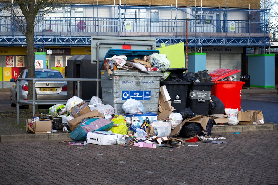 A collection of overflowing waste bins and scattered rubbish situated on a paved area beside a roadside, with a grey recycling container filled with mixed paper, cardboard, and plastic waste in the fo