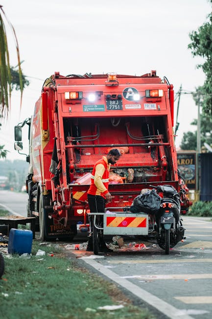 A collection of overflowing waste bins and scattered rubbish situated on a paved area beside a roadside, with a grey recycling container filled with mixed paper, cardboard, and plastic waste in the fo